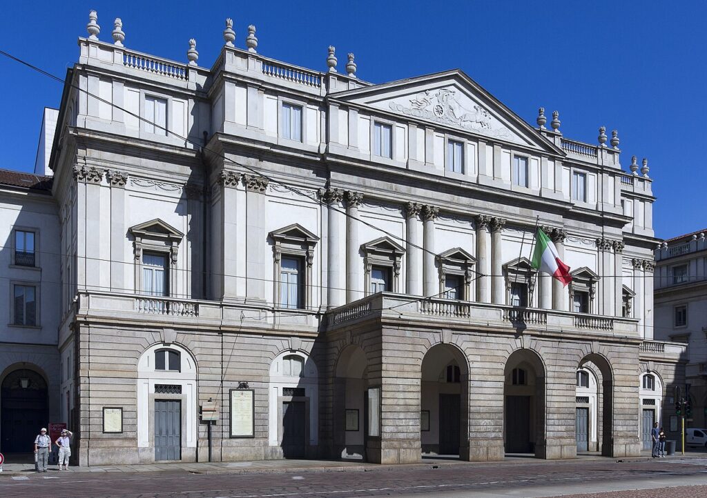 La Scala's pale stone façade beneath a blue sky, with an Italian flag above its entrance.