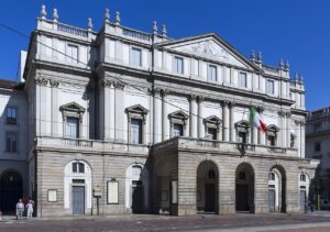 La Scala's pale stone façade beneath a blue sky, with an Italian flag above its entrance.