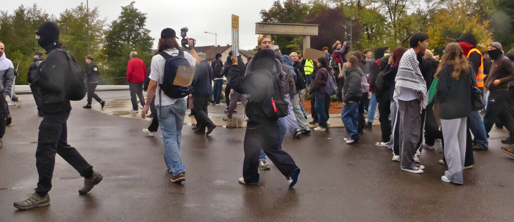 Demonstrators stand and walk on a wet road near the Bavilliers town sign.