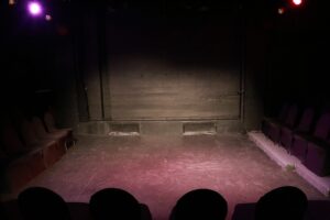 An empty black-box stage with chairs along three sides under colored lights.