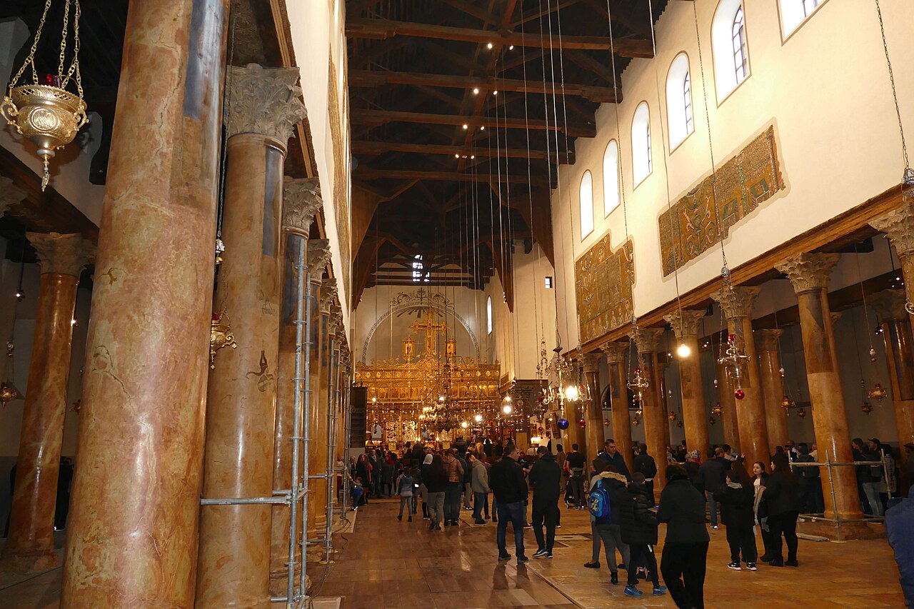 The nave of Bethlehem's Church of the Nativity, with stone columns, hanging lamps and visitors.