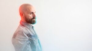 Aiden James in profile, wearing a light denim shirt against a pale background.