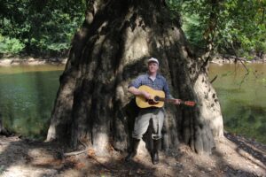 Daniel Bachman holding an acoustic guitar beside a large cypress tree and water.