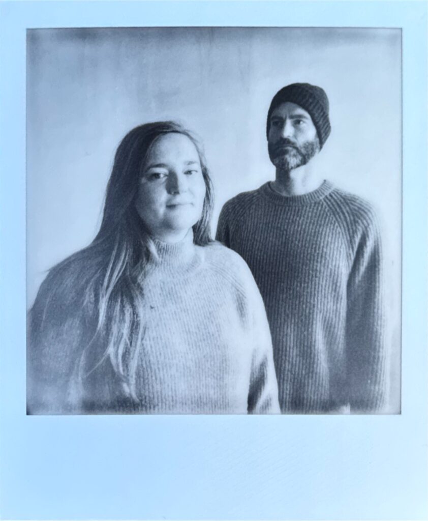 A blue-toned press portrait of Laura James and Oliver Price in knit sweaters.