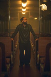 J.MYSTERY standing between seats inside a train carriage.