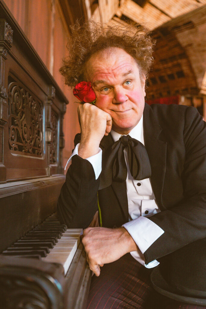 John C. Reilly in a dark suit rests his cheek beside a red rose while seated at a piano.