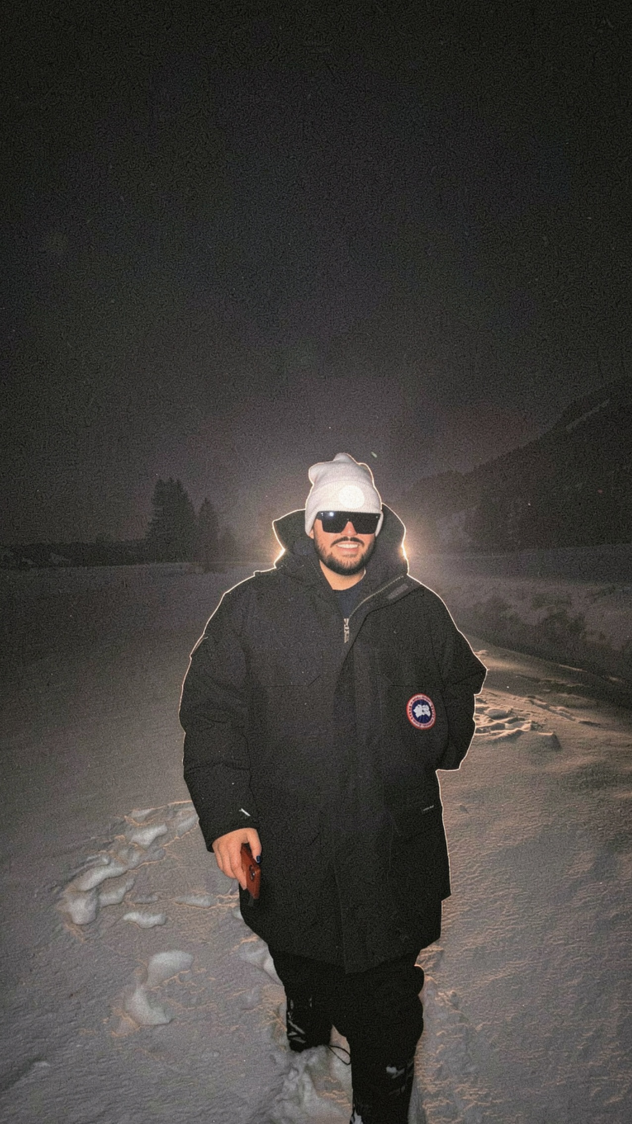 Karim Panzetta wearing a winter coat, knit hat and sunglasses outdoors in the snow at night.