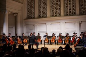 The Suzuki Cello Ensemble performs with a conductor on the Nichols Concert Hall stage.