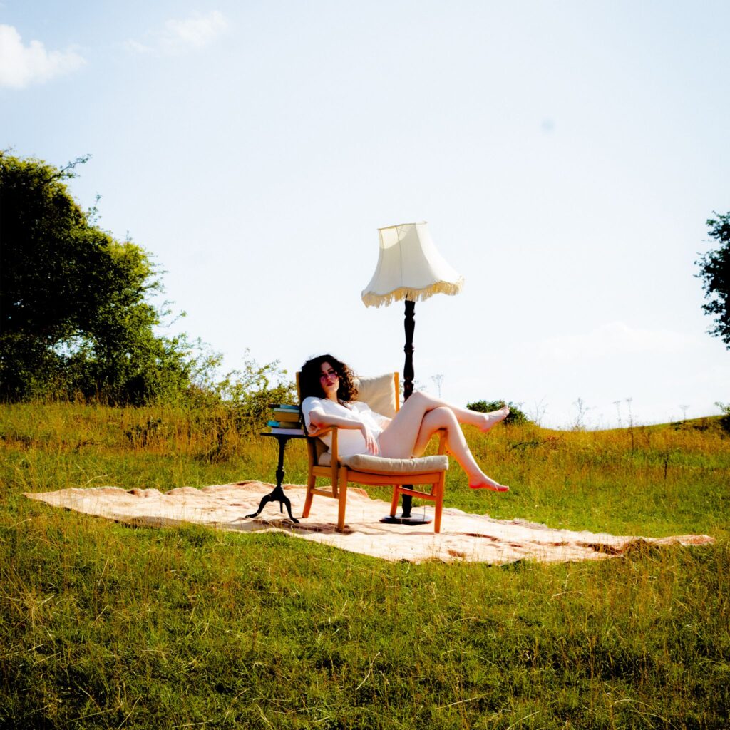 Phoebe Rea sits in an armchair in a grassy field beside a standing lamp and a stack of books.