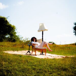 Phoebe Rea sits in an armchair in a grassy field beside a standing lamp and a stack of books.