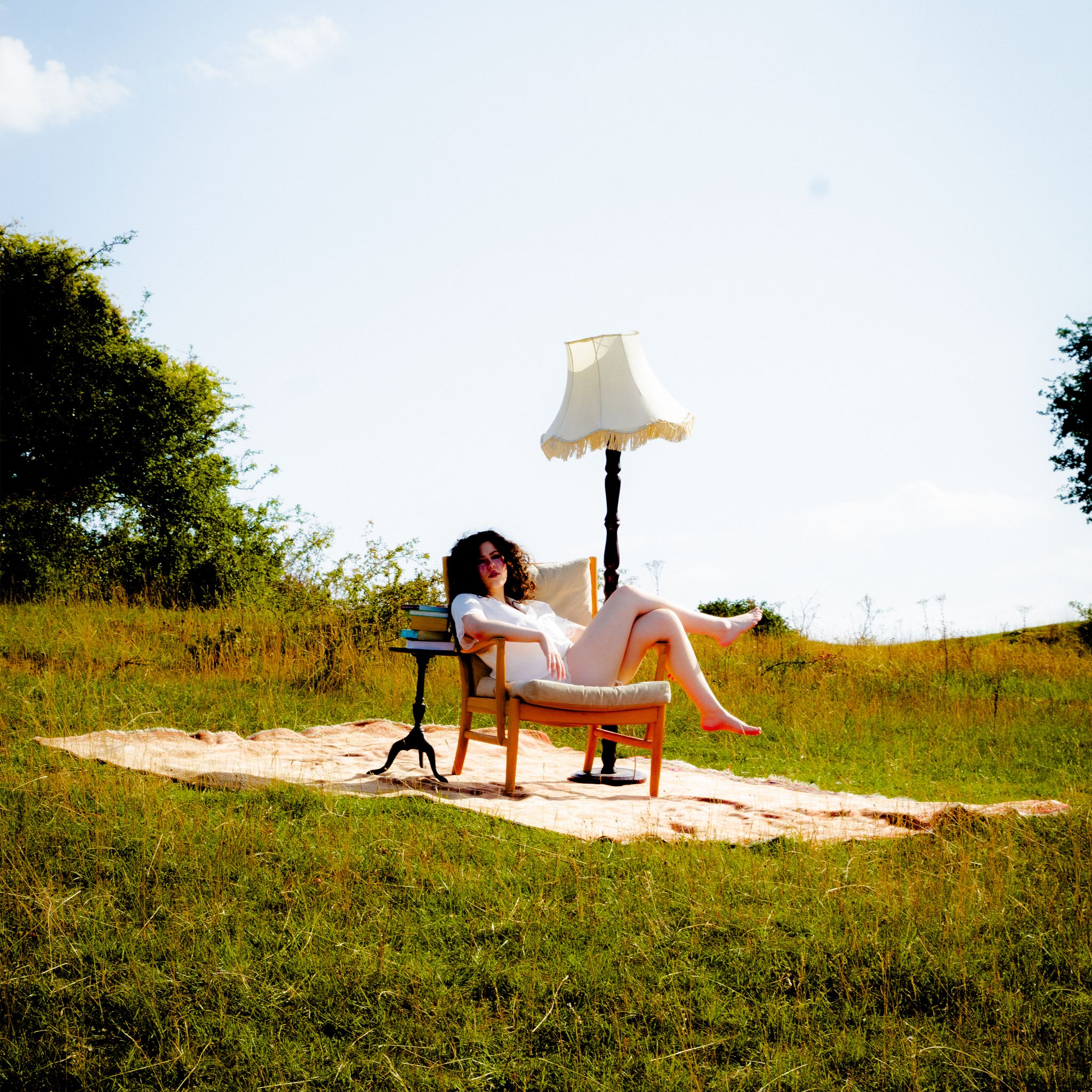 Phoebe Rea sits in an armchair in a grassy field beside a standing lamp and a stack of books.
