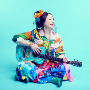 Rachael Sage seated with a decorated acoustic guitar against a turquoise background, wearing a colorful outfit and flowers in her hair.