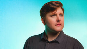 Sacha Mullin in a dark shirt against a turquoise background.