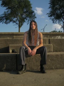 Sen Morimoto seated on concrete steps outdoors, wearing a white sleeveless top, dark trousers and black boots.